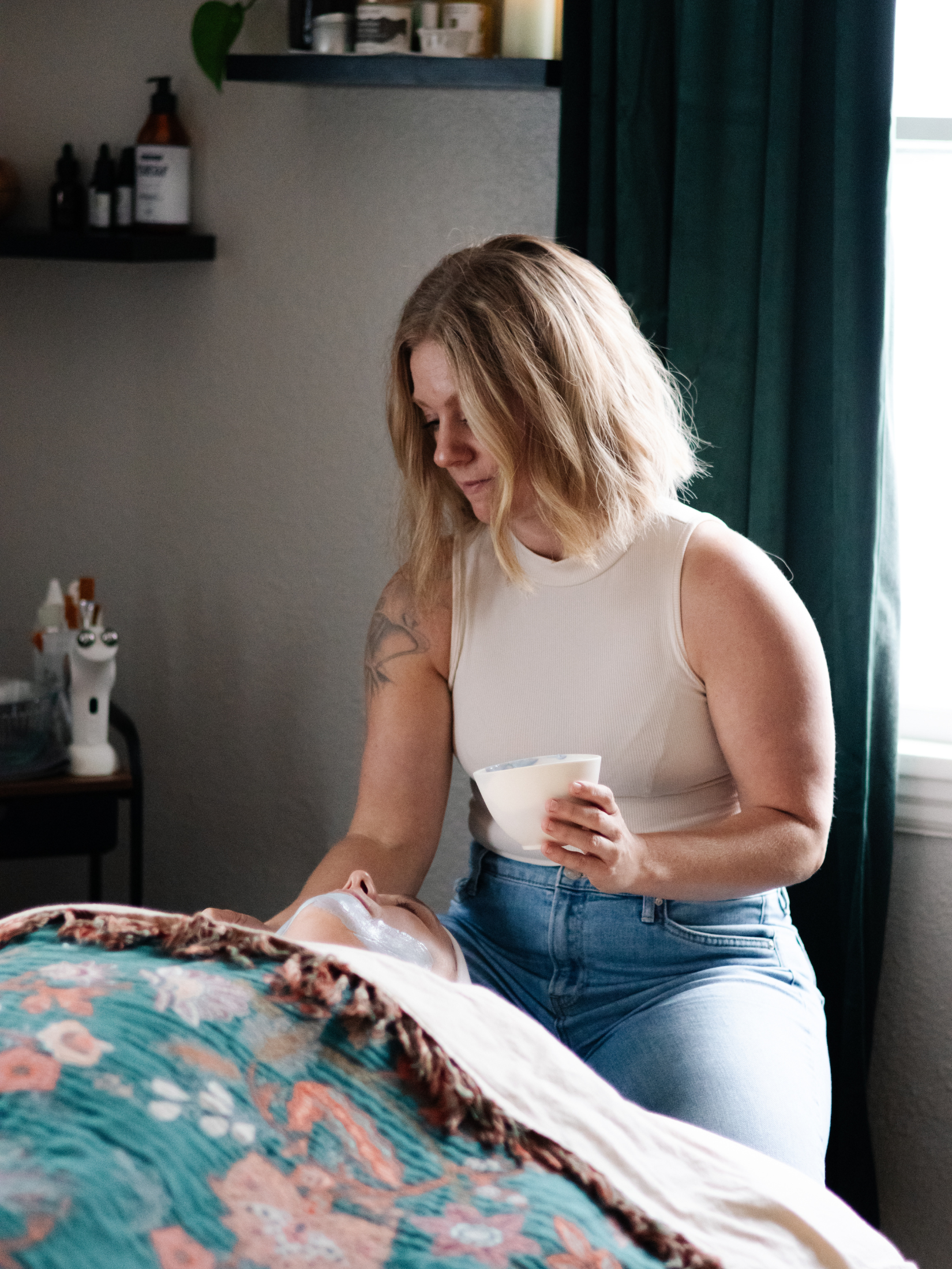 A woman giving a facial to a person lying on a bed, with a bowl in her hand in a cozy bedroom.