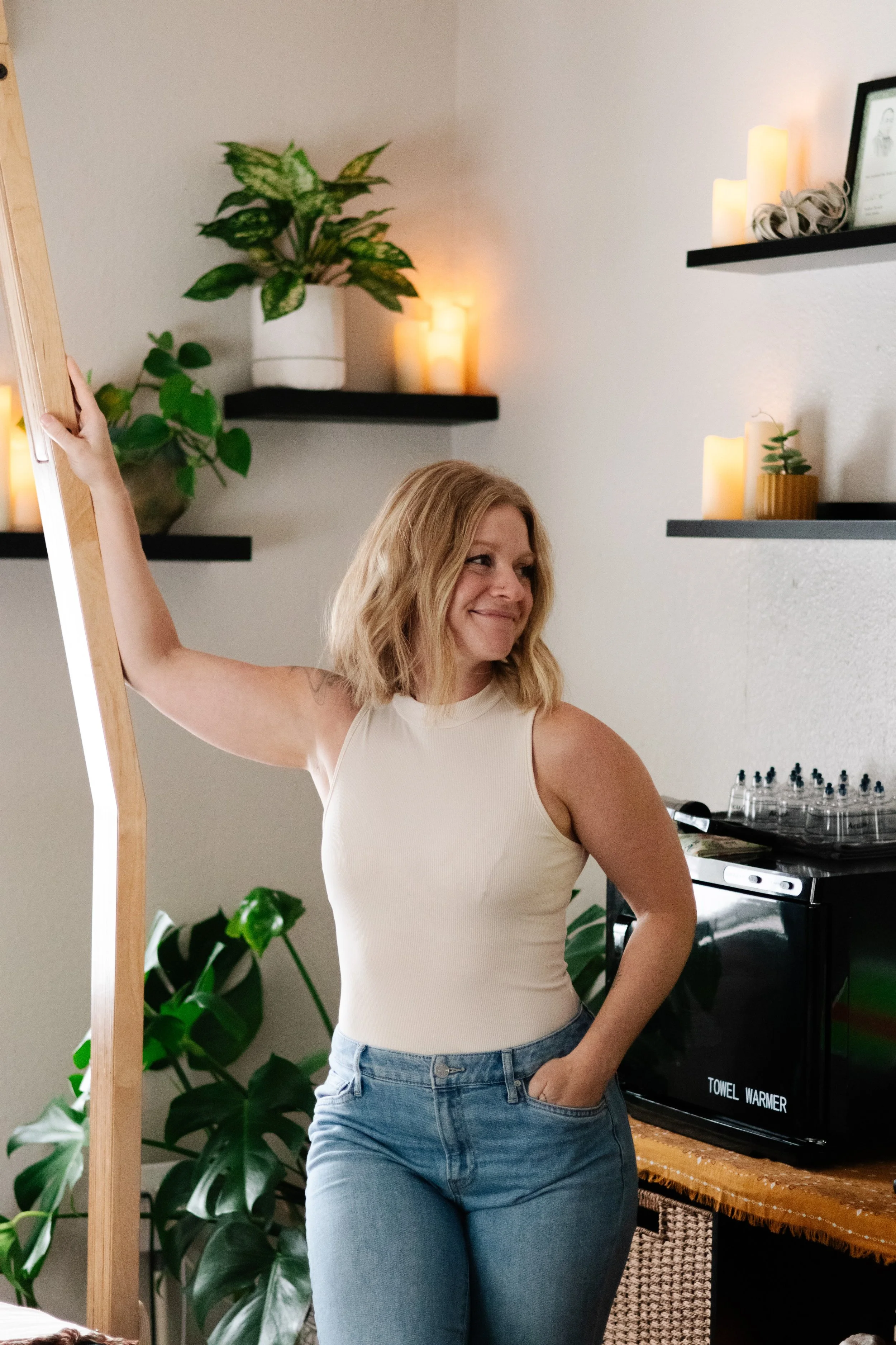 A woman with shoulder-length blonde hair, wearing a sleeveless cream top and blue jeans, stands next to a mirror, smiling and looking to the side in a cozy room with plants, candles, and black shelves.