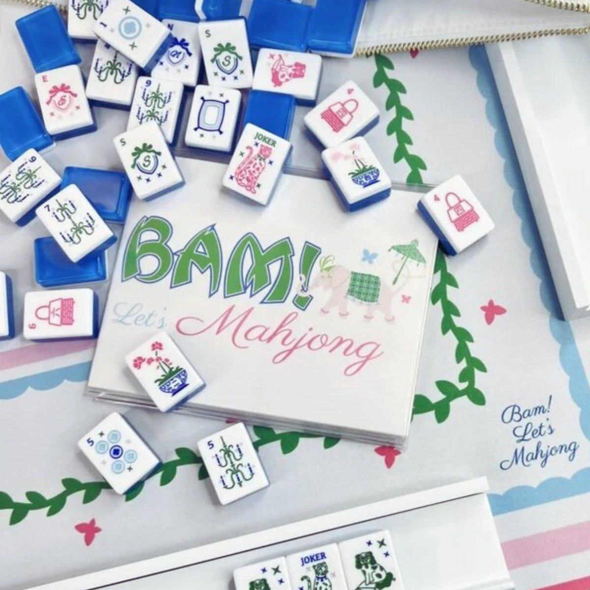 Mahjong game set with tiles scattered on a table, alongside a deck of cards with flowers and umbrellas, on a tablecloth decorated with green and pink patterns and the text 'Bam! Let's Mahjong'.
