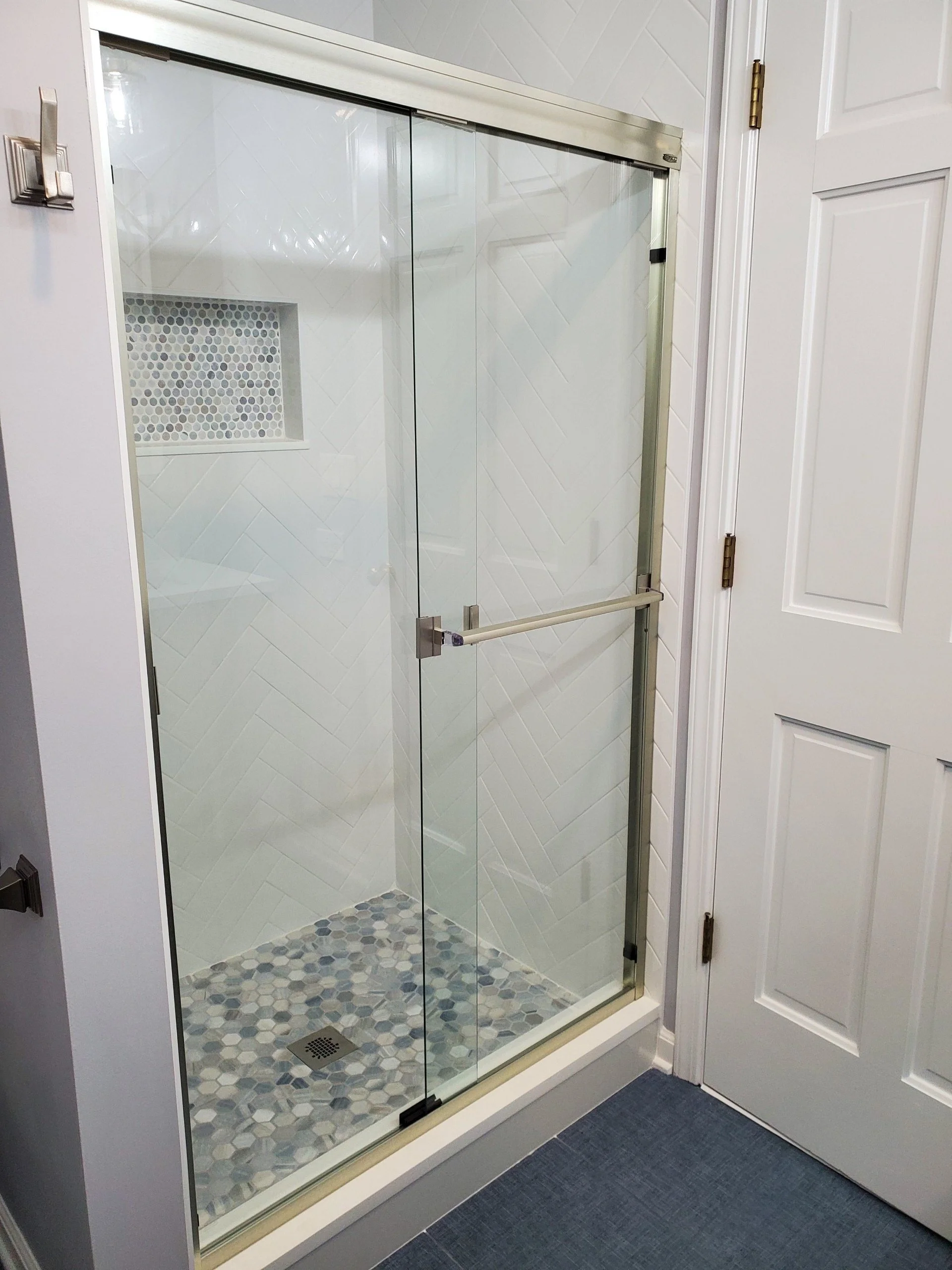 A glass shower enclosure with a metal frame, a mosaic tile floor, and a built-in shelf with small circular tiles inside, next to a white door.