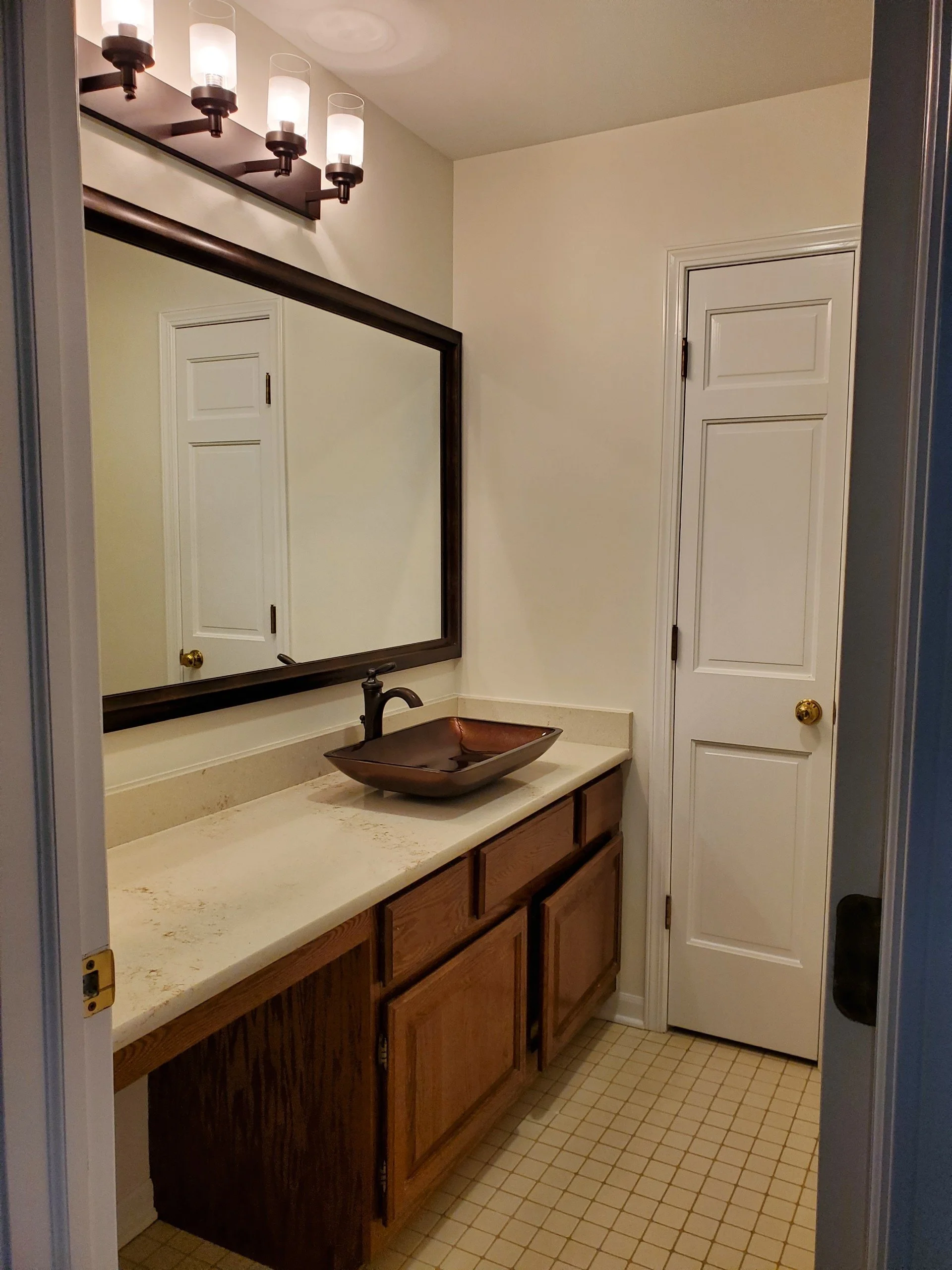 A small bathroom with a wooden vanity, a large mirror, a copper sink, and a light fixture with four pendant lights above the mirror.