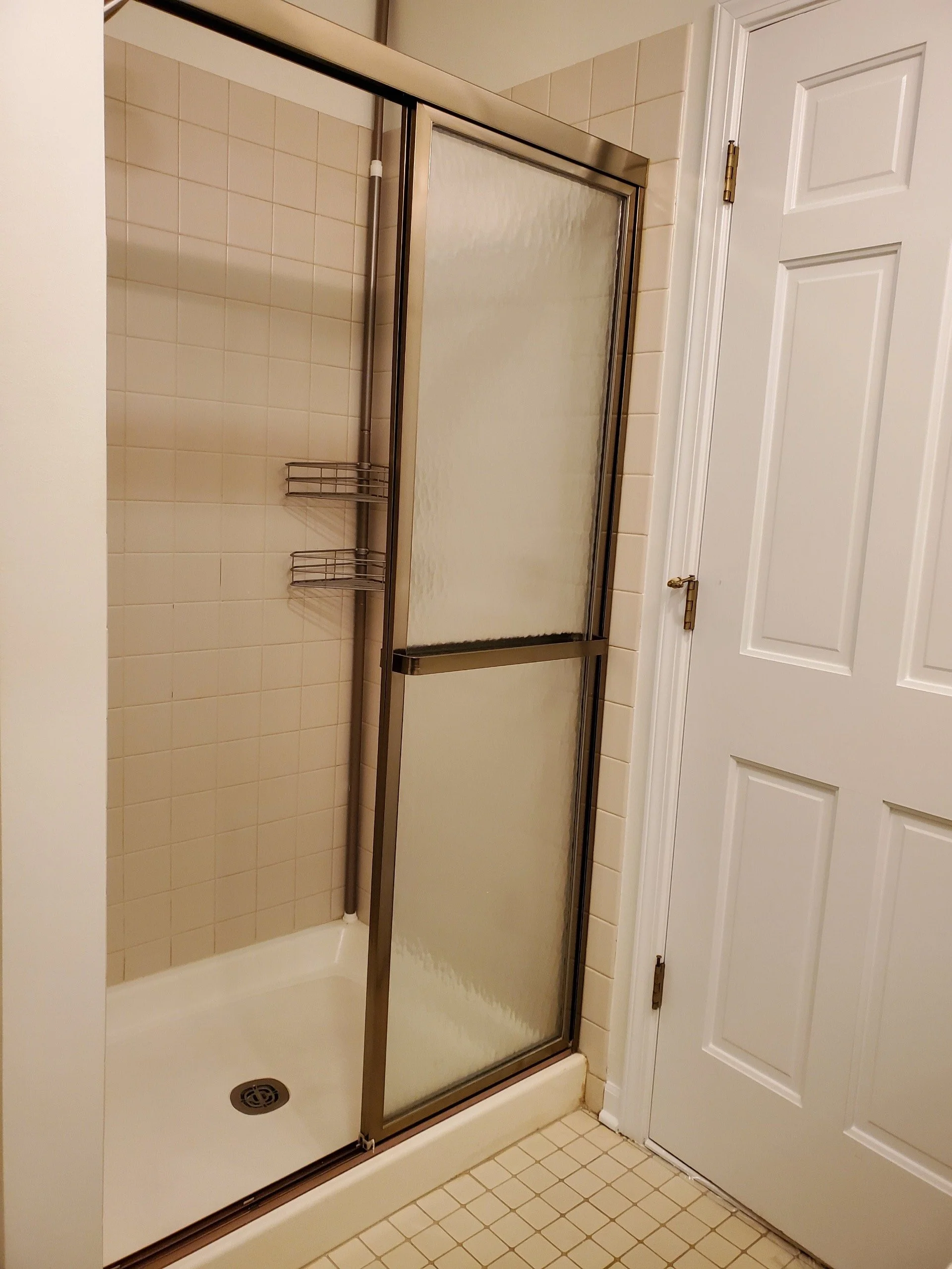 Bathroom shower with frosted glass sliding door, beige tiles, and a small corner shelf. There is a white door next to the shower.