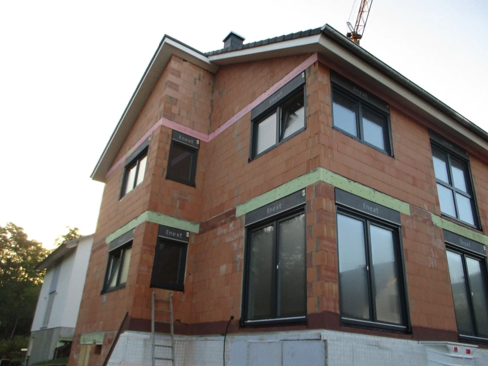 Ein mehrstöckiges Haus im Bau, mit unvollständiger Außenfassade und Fenstern, teilweise mit schwarzem Rahmen installiert, auf einer Baustelle mit Baukran im Hintergrund.