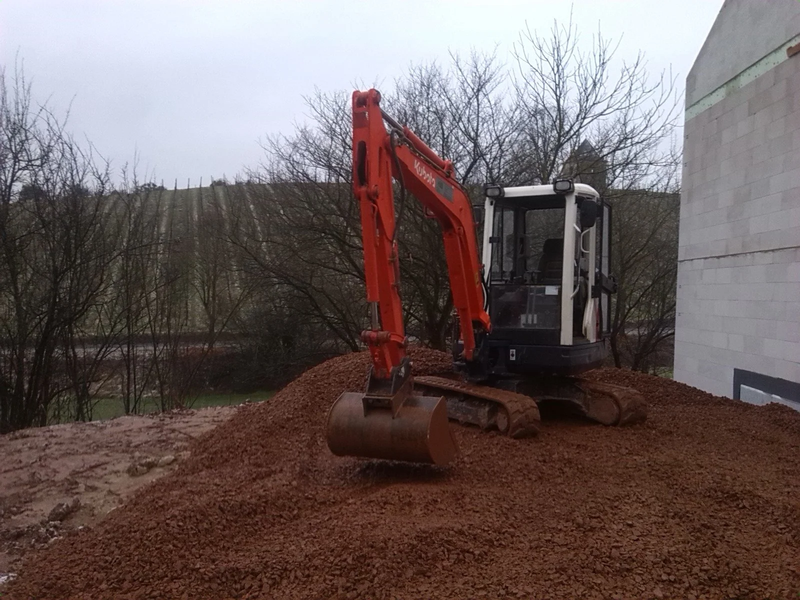 Kleiner Bagger auf einem Erdhaufen neben einem weißen Gebäude, im Hintergrund Bäume und ein bewölkter Himmel.