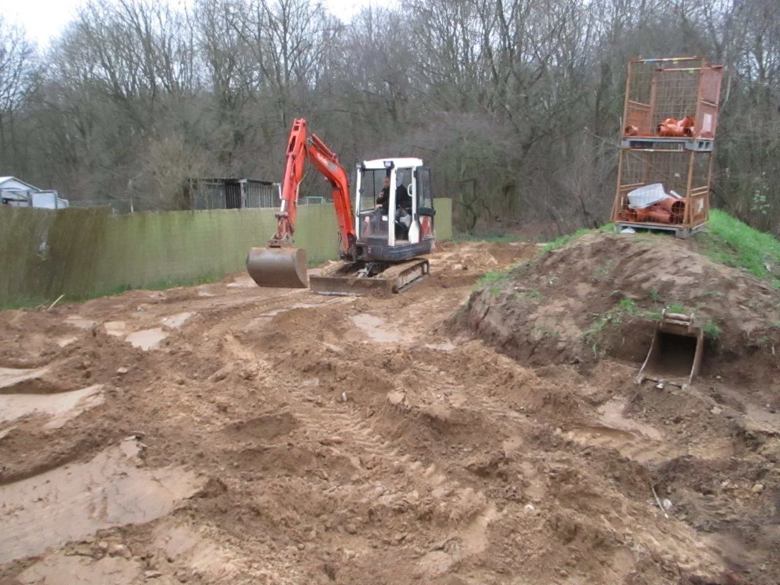 Baugrund mit Minibagger und Bauschutt im Freien, im Hintergrund Bäume und eine grüne Mauer.