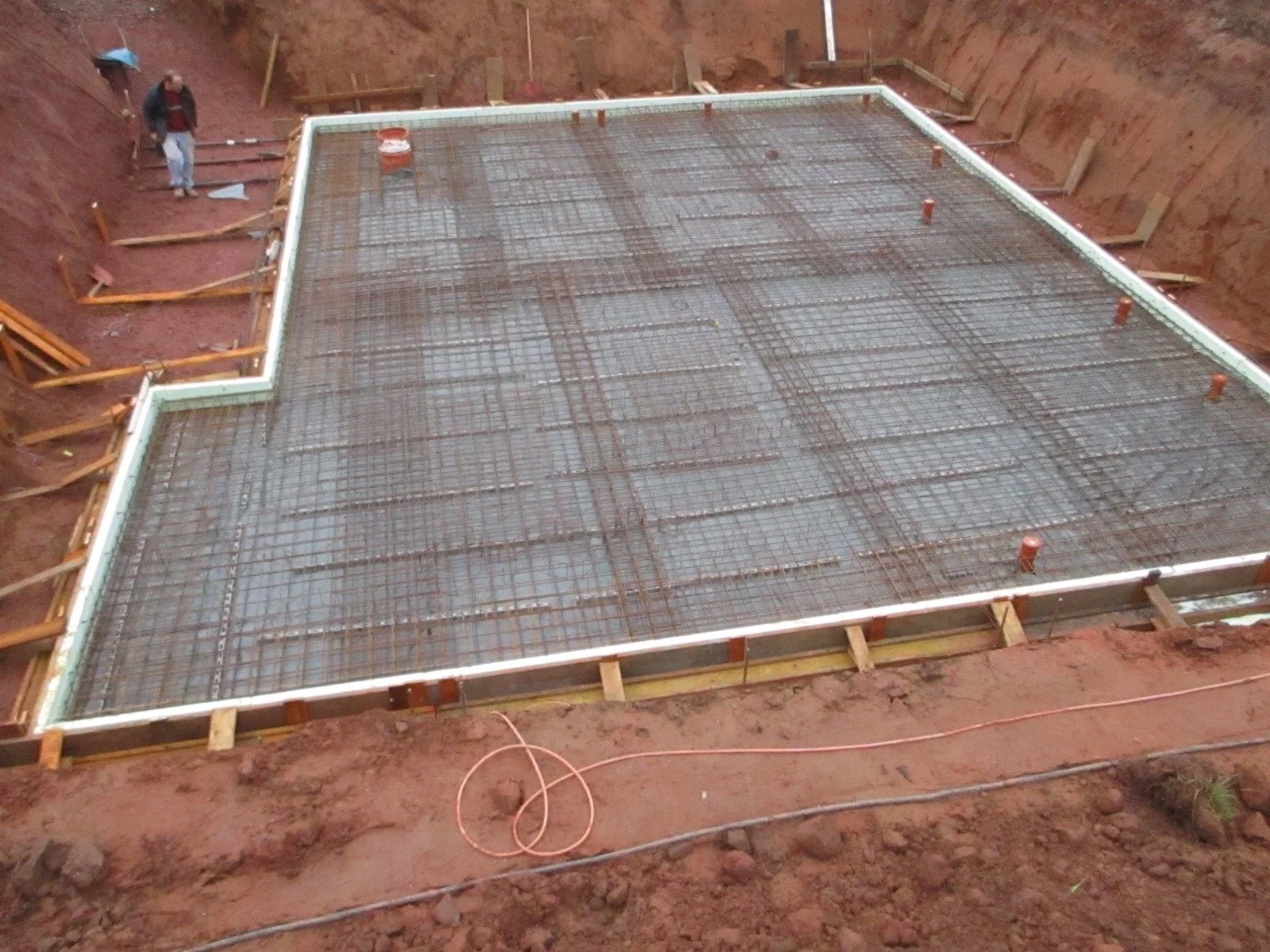 Fundament eines Gebäudes mit Stahlbewehrung in einem Erdloch, Bauarbeiter im Hintergrund.