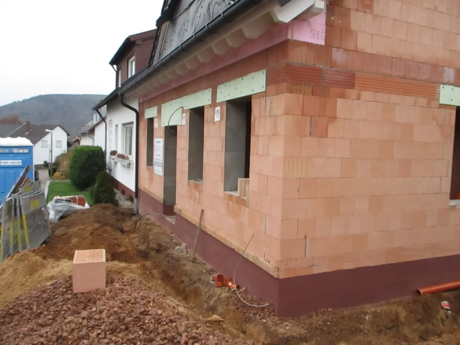 Rohbau eines Hauses mit Ziegelwänden, Fensteröffnungen und Baugerüsten, im Vordergrund Erdarbeiten.