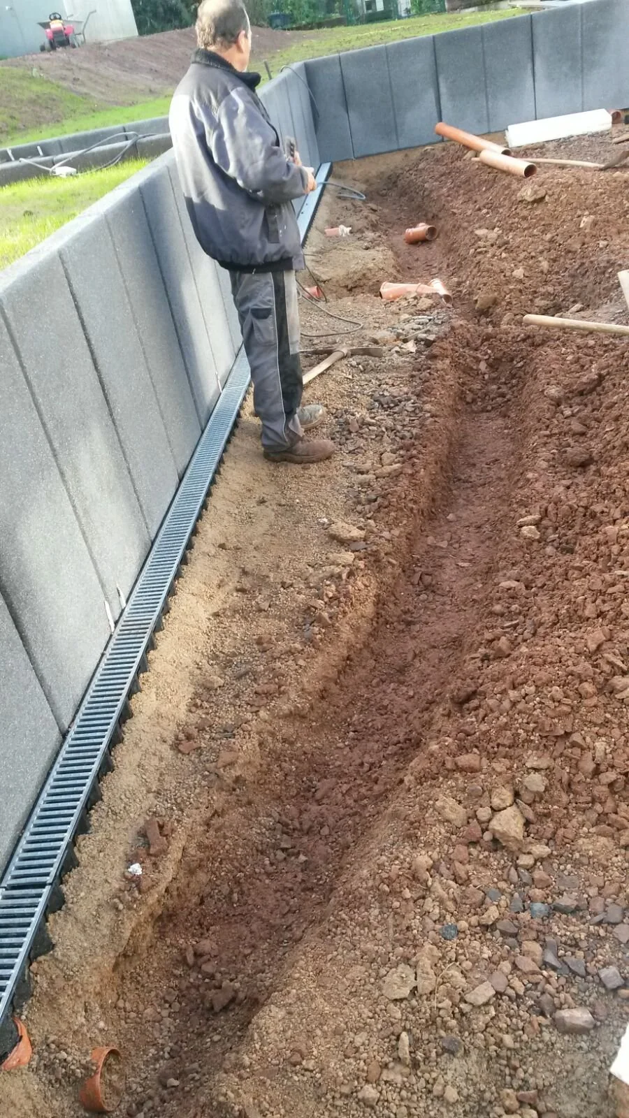 Ein Mann steht neben einer Baugrube mit rotem Erdreich, mehreren Rohren und einer Regenrinne, auf einer Baustelle im Freien.