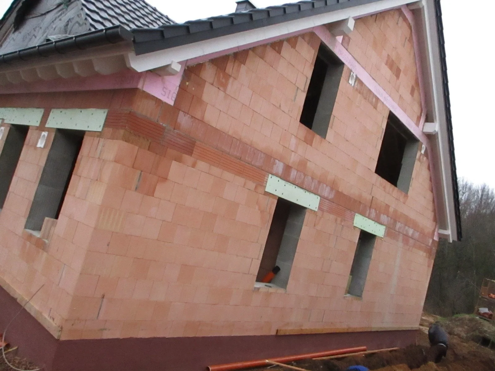 Rohbau eines Hauses mit Sichtmauerwerk aus roten Ziegelsteinen, offene Fensteröffnungen, im Hintergrund Bäume und Baugeräte.