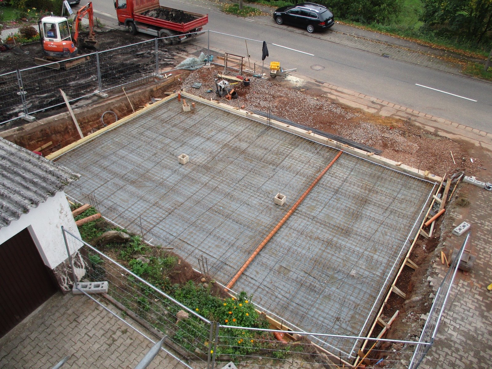 Baustelle mit vorbereiteter Betonboden, Baustellenzaun, Baugeräte und Fahrzeuge in einer städtischen Gegend.