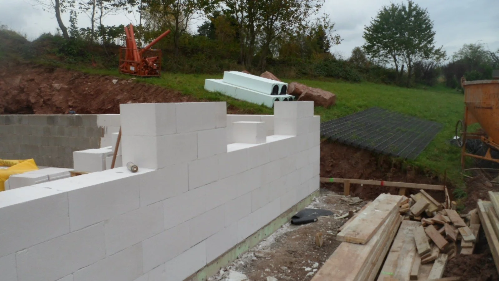 Baustelle mit weißen Ziegelwänden, Holzbohlen, Baumaterialien und Baugeräten im Freien, umgeben von Bäumen und Erde, bei bewölktem Himmel.