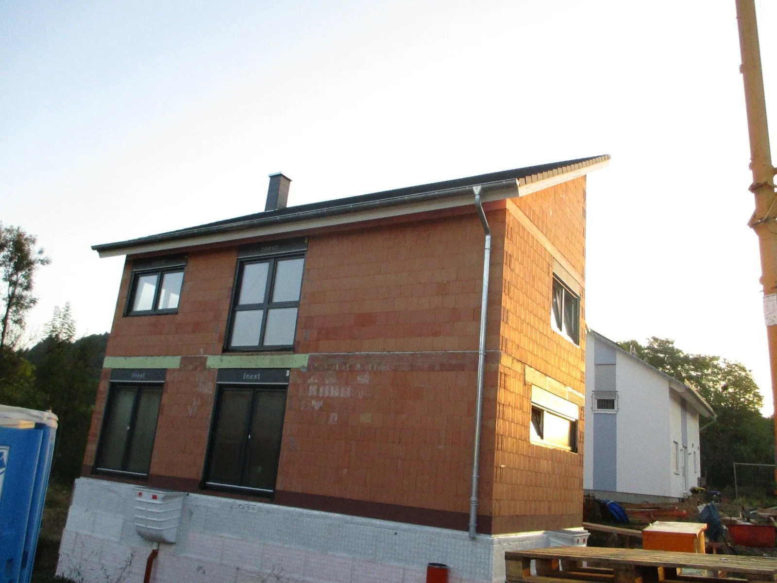 Ein zweistöckiges Haus unter Bau mit rotem Backsteinmauerwerk, mehreren Fenstern und einem grauen Dach, Sonnenlicht spiegelt sich auf den Glasflächen.