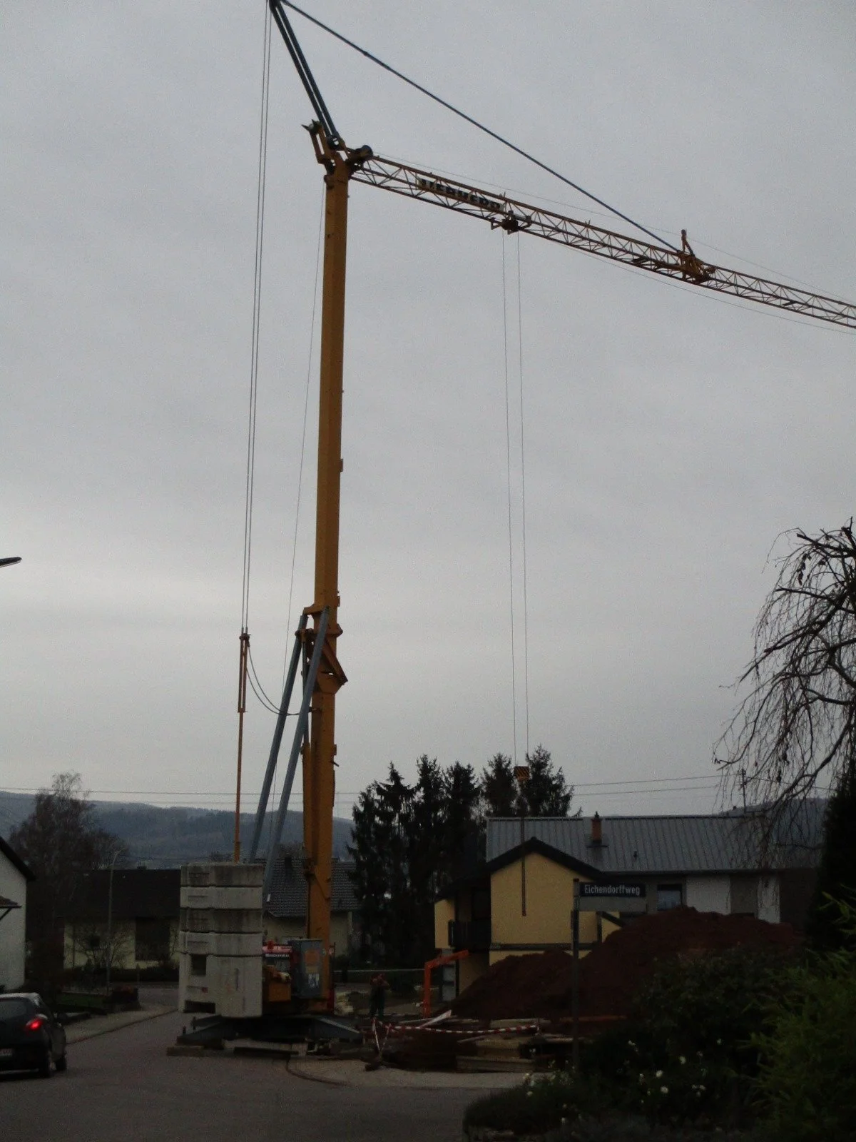 Ein Baukran steht auf einer Straße, mit umstehenden Häusern und einem Hügel im Hintergrund, bei bewölktem Himmel.