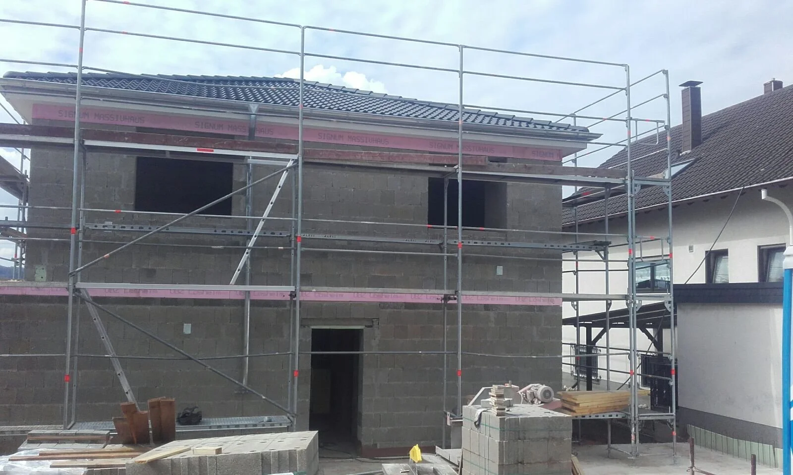 Baustelle eines Mehrfamilienhauses mit Gerüsten, unfertiges Gebäude aus Ziegelsteinen, Bauholz, Werkzeuge und Baumaterialien im Vordergrund, neben einem fertiggestellten Haus mit dach und Schornstein