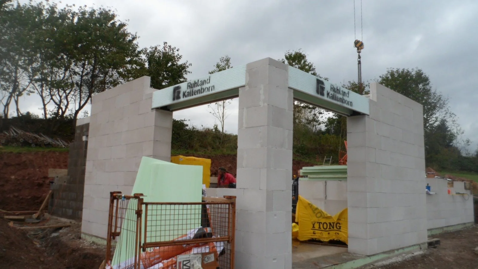 Baustelle mit weißen Ziegelwänden und Bauarbeiten, umgeben von Bäumen, mit einem Baukran im Hintergrund, es ist bewölkt.