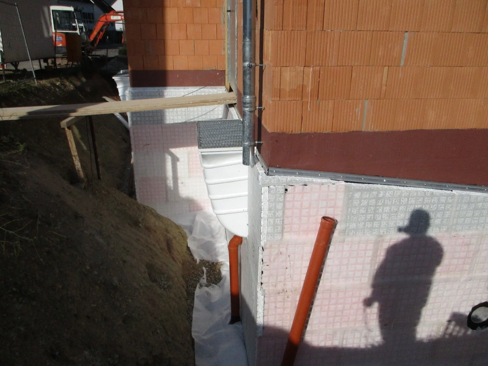 Baustelle mit rotem Mauerwerk, Rohren und Baugerüsten, Schatten einer Person mit Helm im Vordergrund.