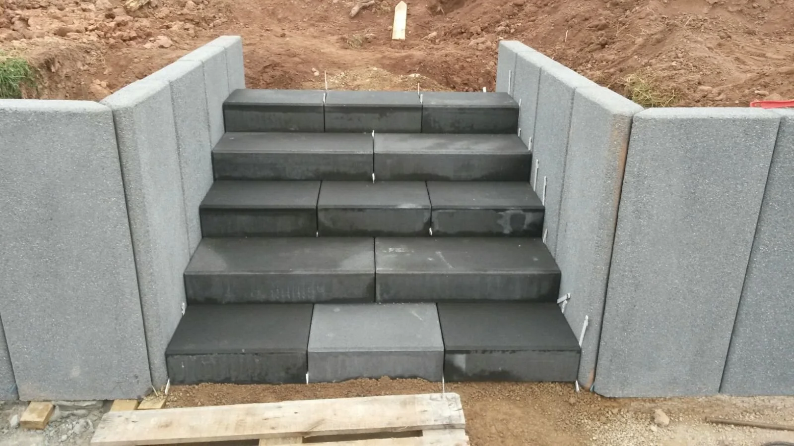 Steintreppe mit schwarzen Fliesen zwischen zwei Zäunen aus grauem Beton auf einem Baustellengelände.