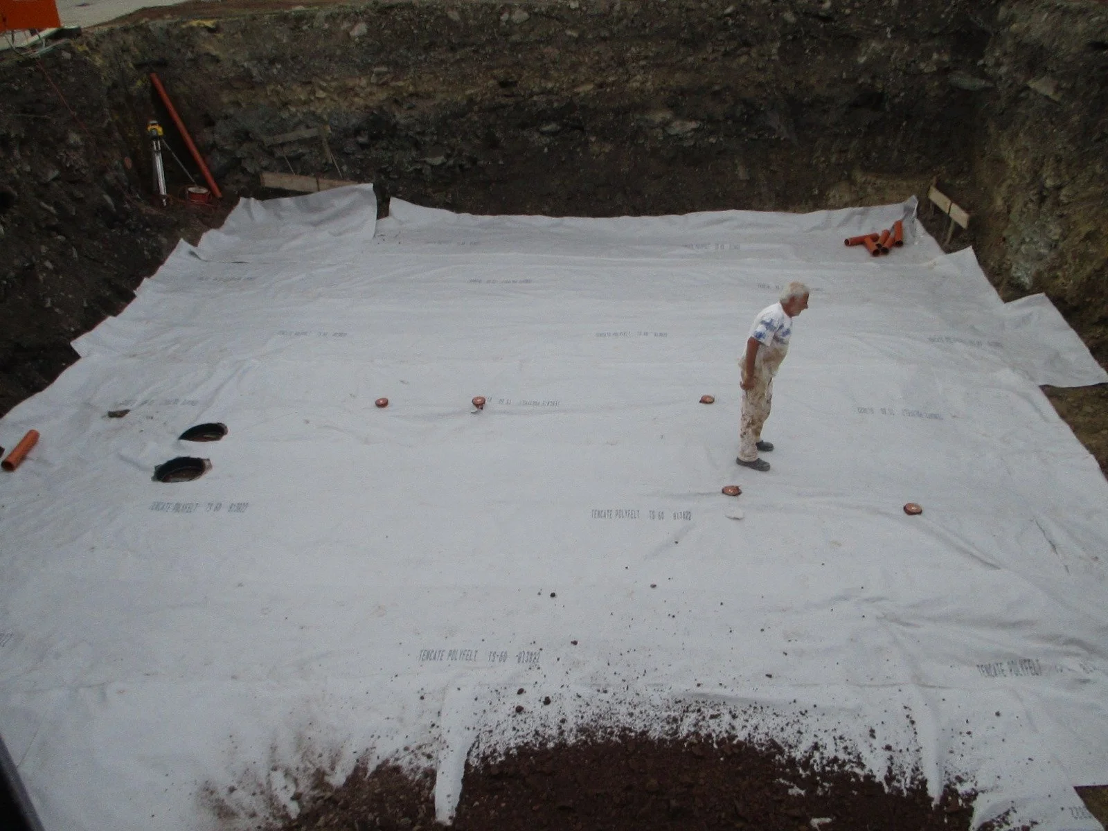 Bauarbeiter auf einer Baustelle, Boden wird mit Isoliermaterial bedeckt, umgeben von Erdarbeiten und Rohren.