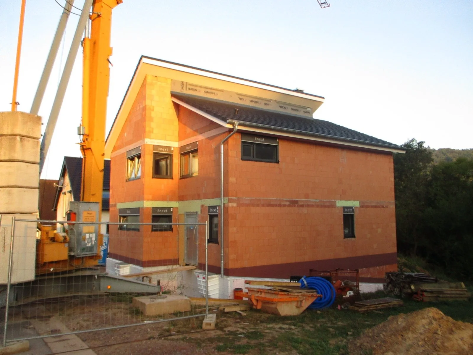 Ein zweistöckiges Haus wird gebaut, mit sichtbaren roten Ziegelwänden und dunklen Fenstern, auf einer Baustelle mit Baugeräten, Rohren und Baumaterialien im Vordergrund.