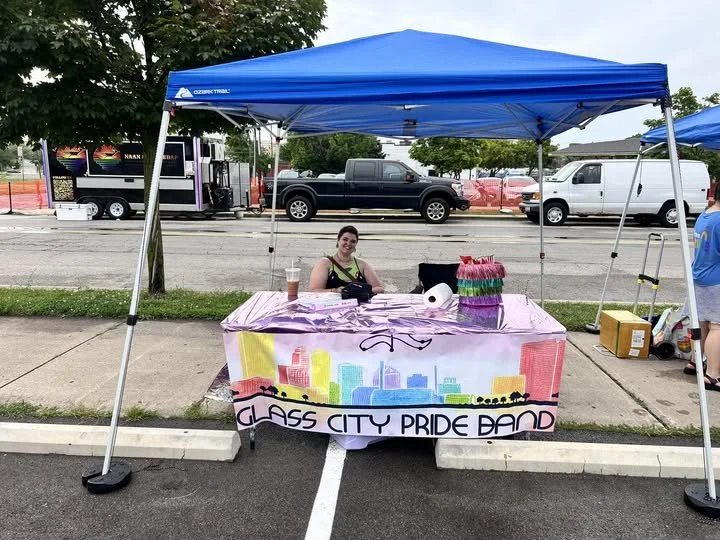 A person sitting at a table under a blue canopy at a pride event. The table has a colorful banner with a city skyline that reads 'Glass City Pride Band.' There are rainbow-colored decorations on the table.