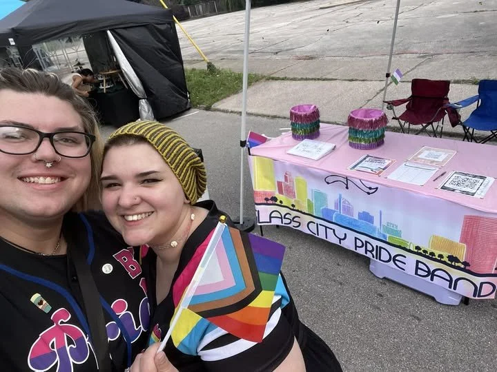 Two people smiling at the pride event, one holding a rainbow pride flag, with a table titled "Glass City Pride Band" and informational materials behind them.