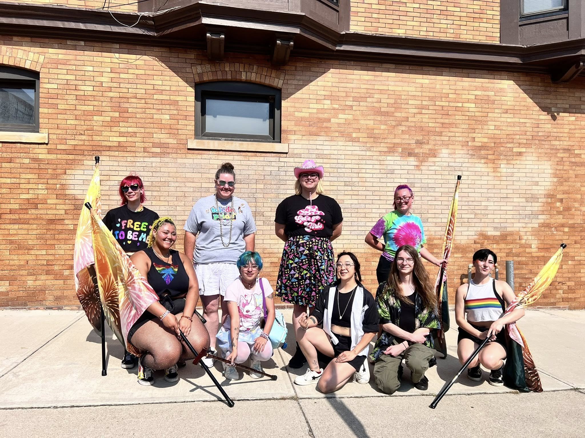 A group of eleven diverse people celebrating Pride, holding rainbow flags and wearing colorful clothing, standing and kneeling in front of a brick wall on a sunny day.