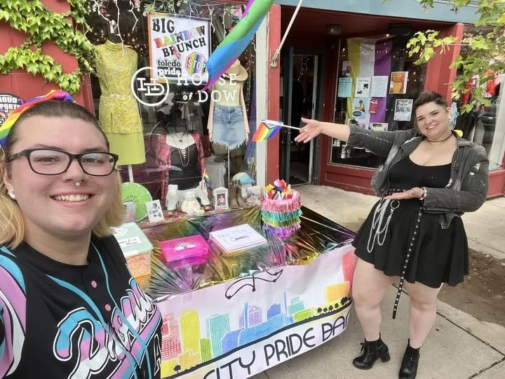 Two smiling women at a pride event, one taking a selfie with a table displaying pride-themed items like rainbow flags and decorations outside a building. The background has a cityscape banner and posters.