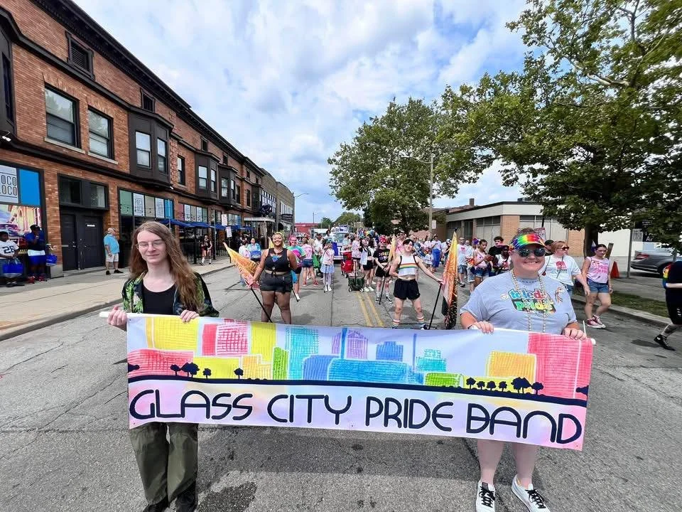 People participating in a pride parade, holding a colorful banner that reads 'Glass City Pride Band' with a rainbow-colored cityscape illustration, on a city street lined with brick buildings and trees.