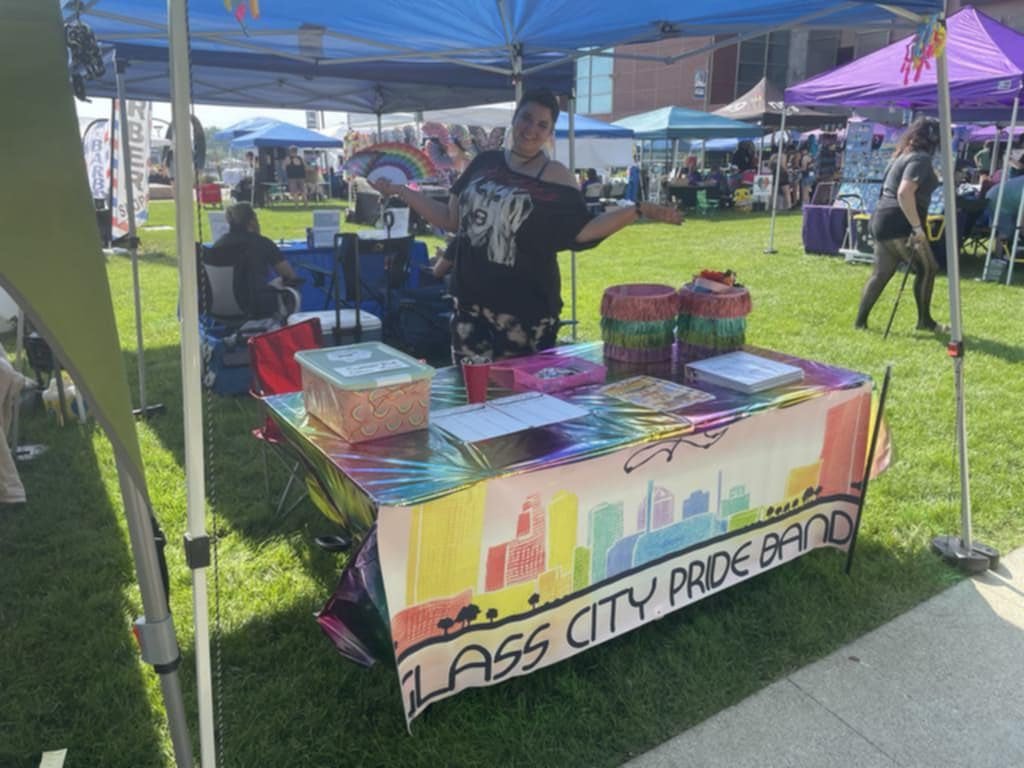 A woman standing behind a booth at an outdoor event, displaying rainbow-colored items, with a rainbow and cityscape illustration on the tablecloth that reads 'Glass City Pride Band"