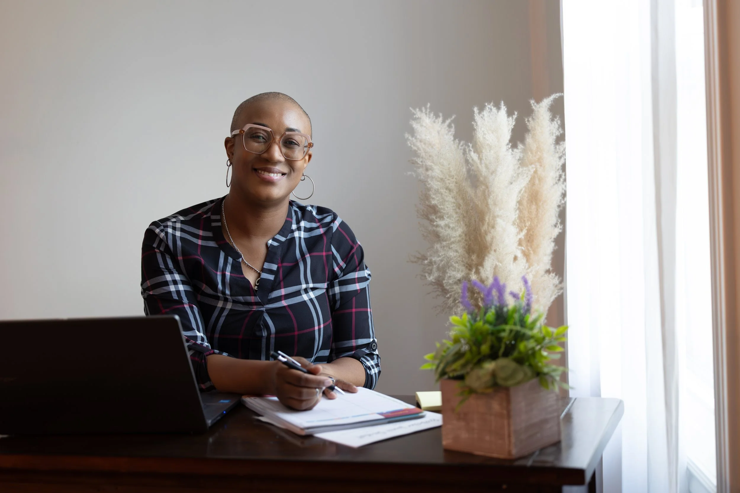 A smiling woman with glasses and earrings sitting at a dark wooden desk, holding a pen, with a laptop and notebooks in front of her, next to a potted plant with purple flowers and beige pampas grass, near a window with white curtains.