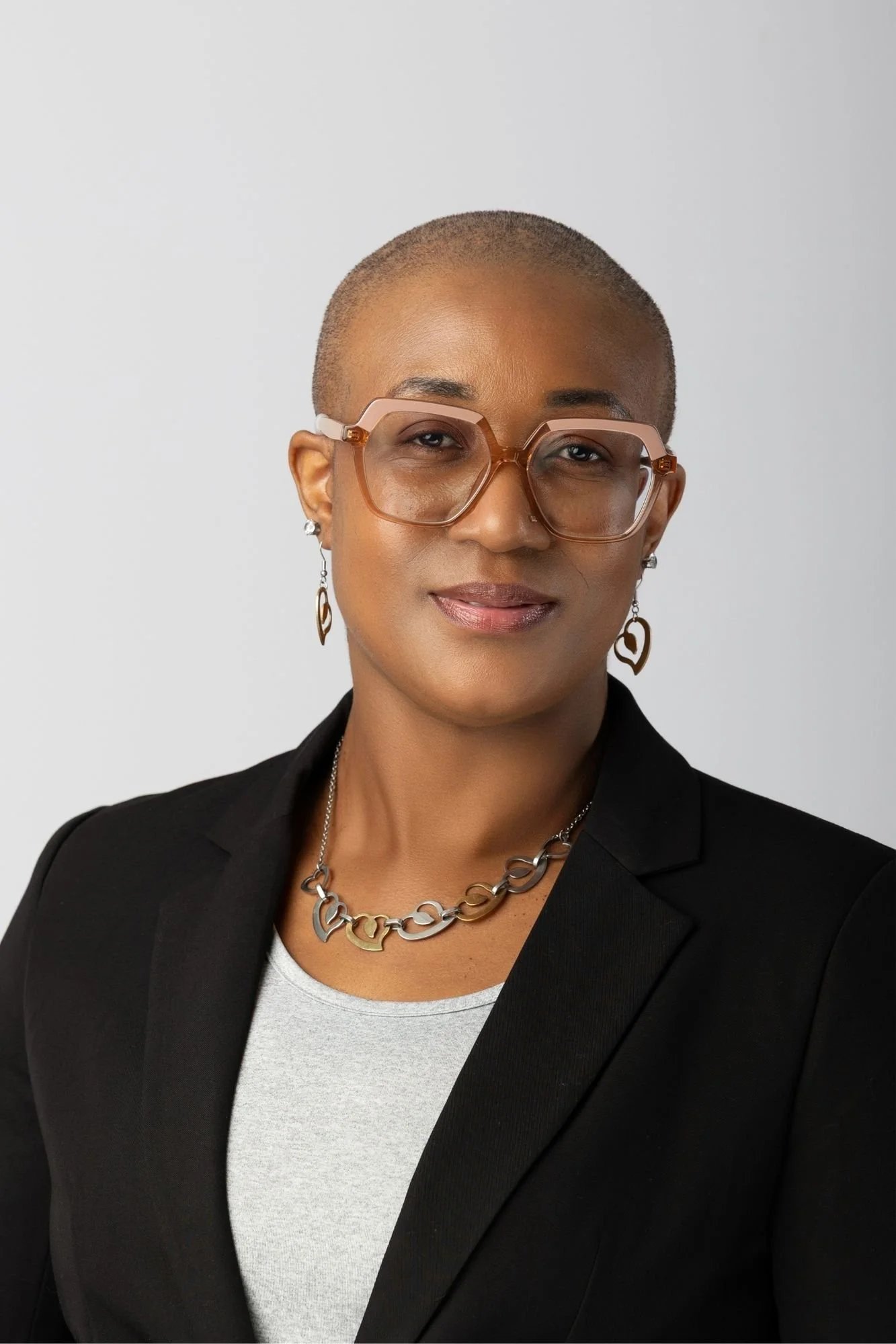 Professional Headshot of a confident Tamica Daley with a shaved head, wearing glasses, hoop earrings, a silver and gold chain necklace, a grey shirt, and a black blazer against a plain light background representing leadership presence.