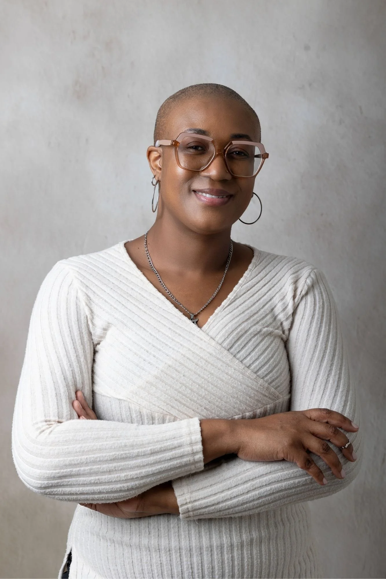 Tamica Daley with glasses and earrings smiling, with arms crossed, wearing a cream-colored ribbed sweater against a plain background.