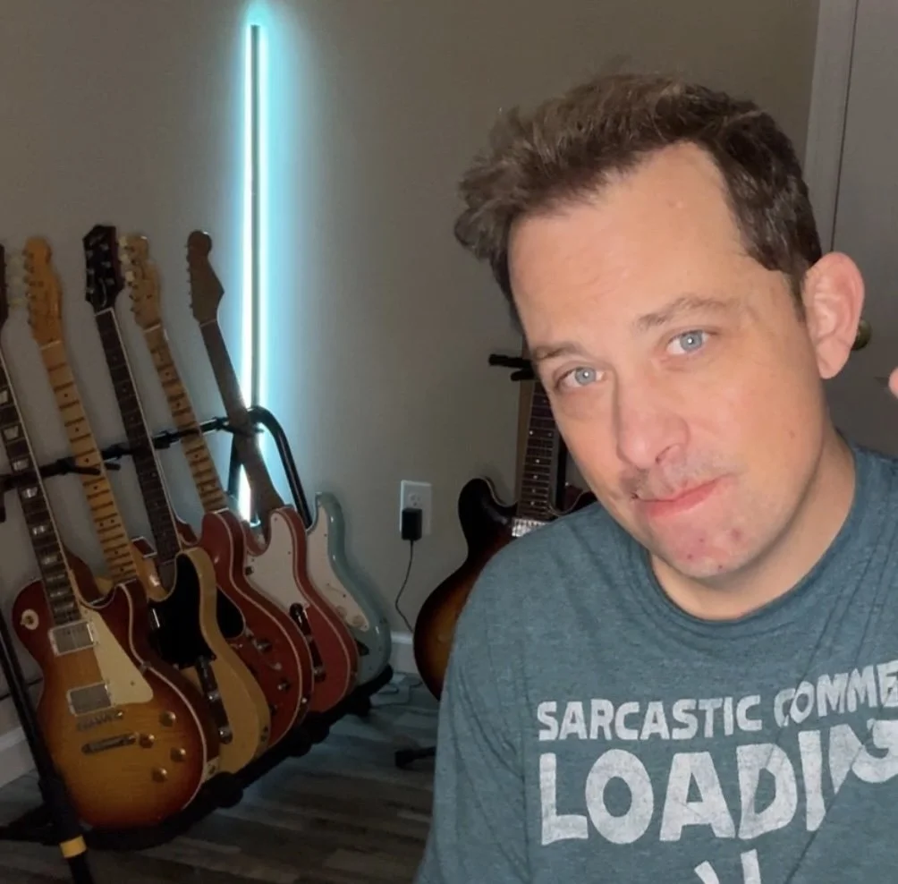 A man with short brown hair and blue eyes taking a selfie in front of six guitars on a stand, with a black and brown electric guitar on the wall behind him.
