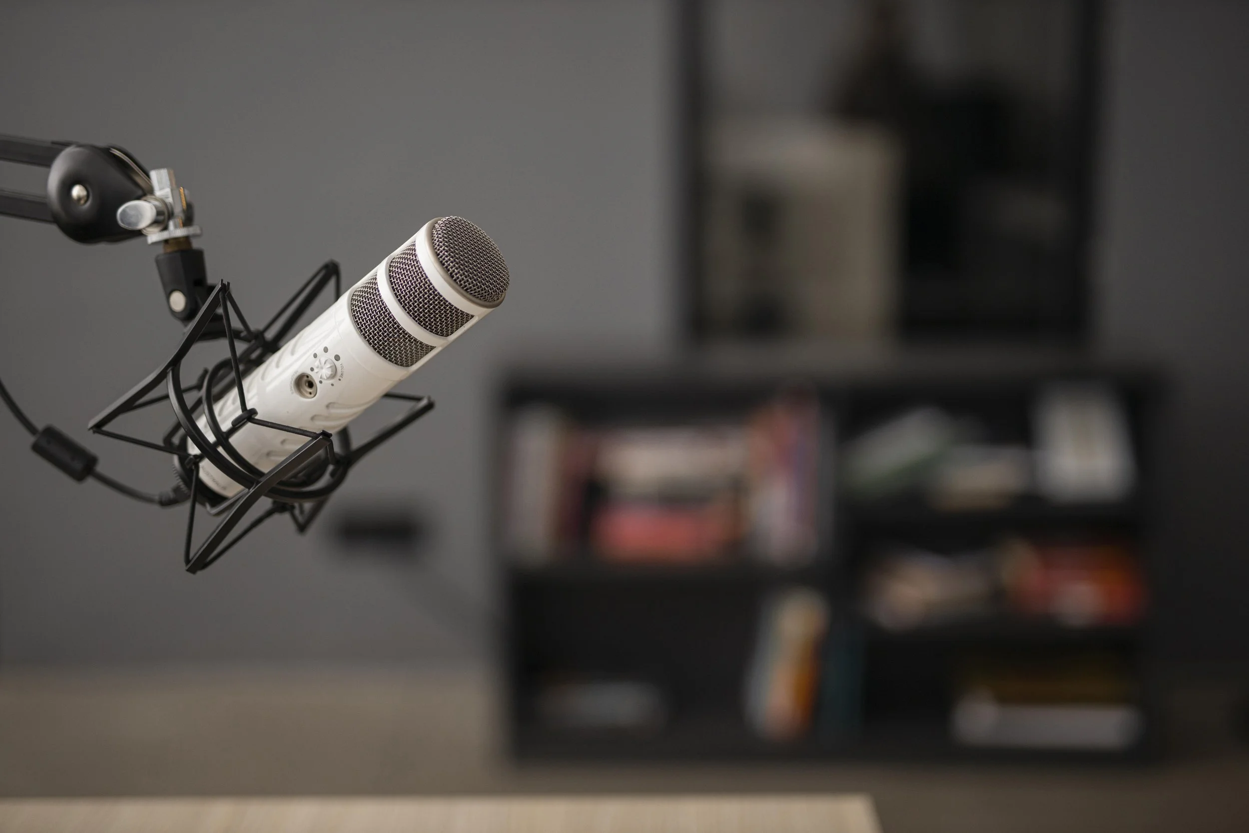 A white microphone mounted on a black shock mount in a recording studio with shelves filled with books and items in the background.
