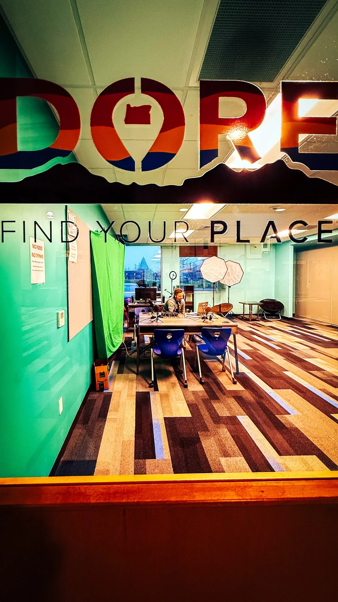 View through glass window showing a modern office or studio with a person working at a desk, green wall, and professional lighting equipment, with the words 'Find Your Place' on the glass.