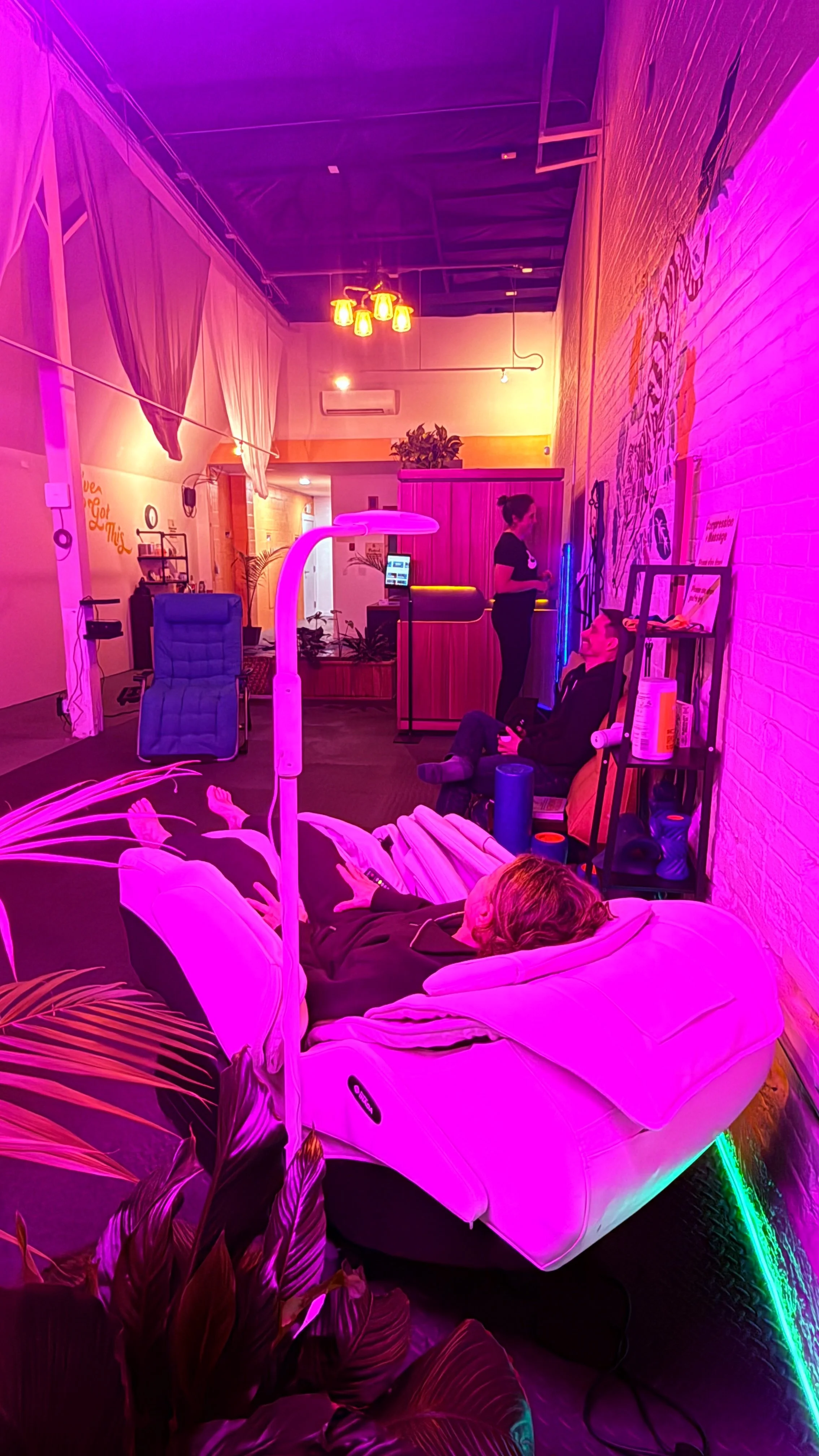Interior of a modern, brightly lit wellness or massage studio with colorful pink and purple lighting, massage beds, a reclining chair, and a staff member standing near a reception desk.