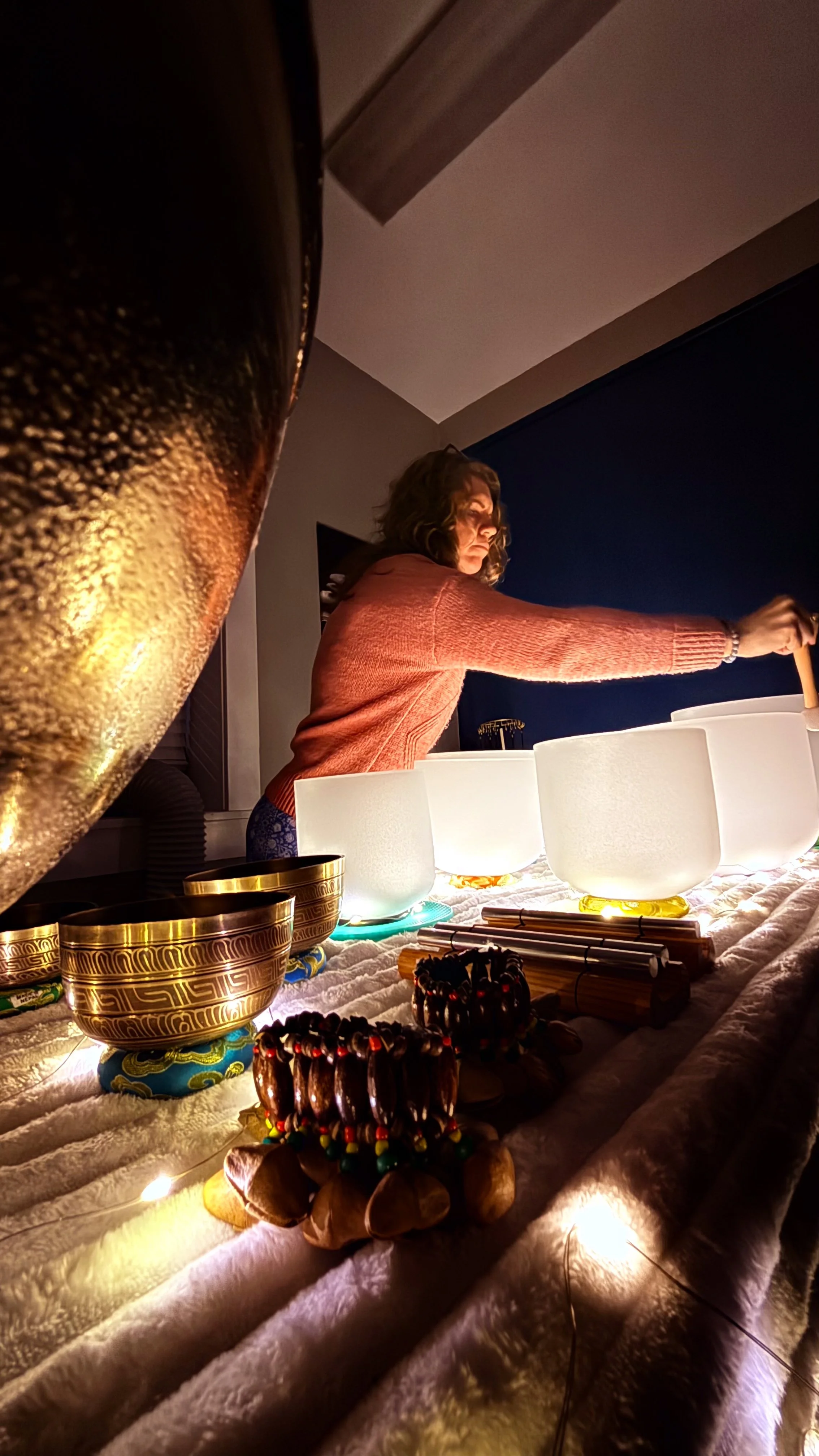 A woman performing a musical ritual with crystal singing bowls and decorated drums on a table. She is wearing a pink sweater and is focused on playing the bowls with a mallet.