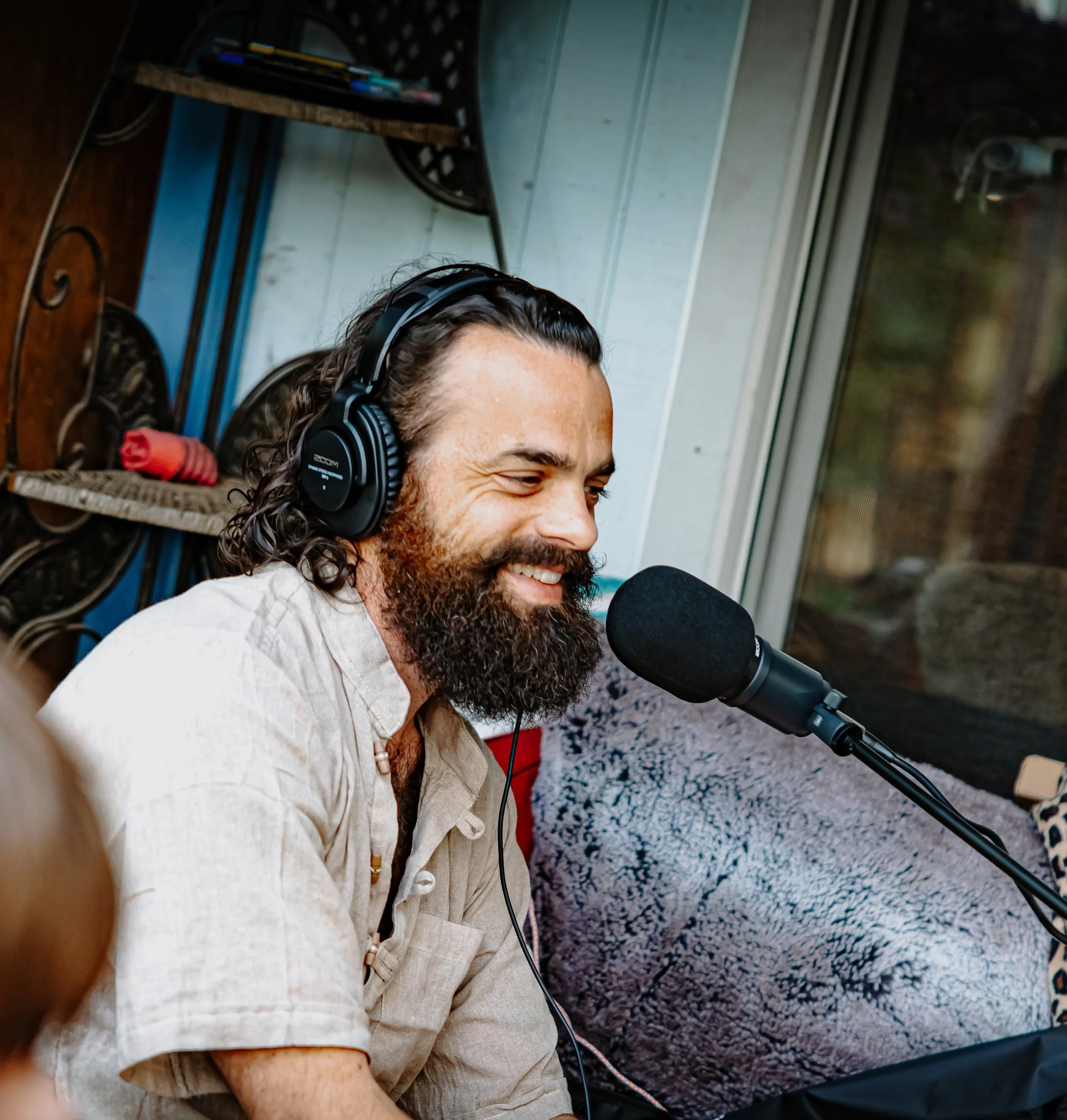 A man with long dark wavy hair and a thick beard wearing headphones and smiling while speaking into a microphone.