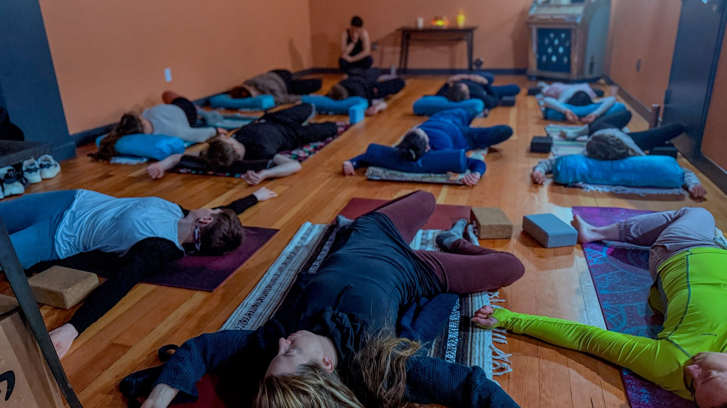 People participating in a group yoga or meditation class, lying on mats on the wooden floor in a room.