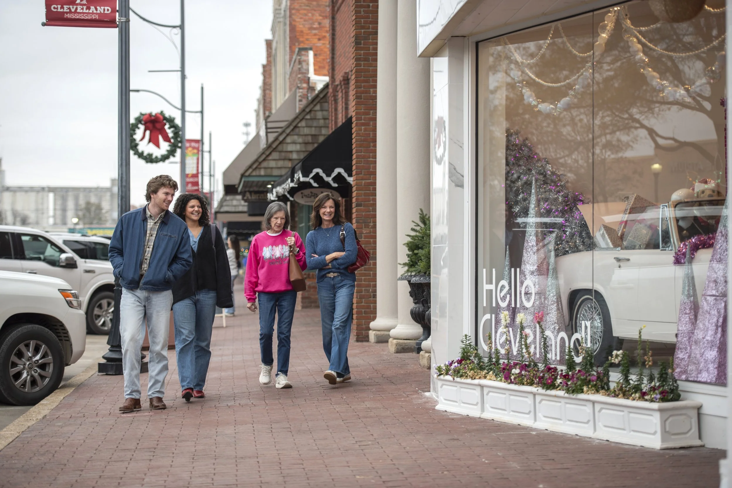 Four women walking along a brick sidewalk in a small downtown area decorated for Christmas, with a storefront window displaying holiday decorations and a sign that says 'Hello Cleveland'.