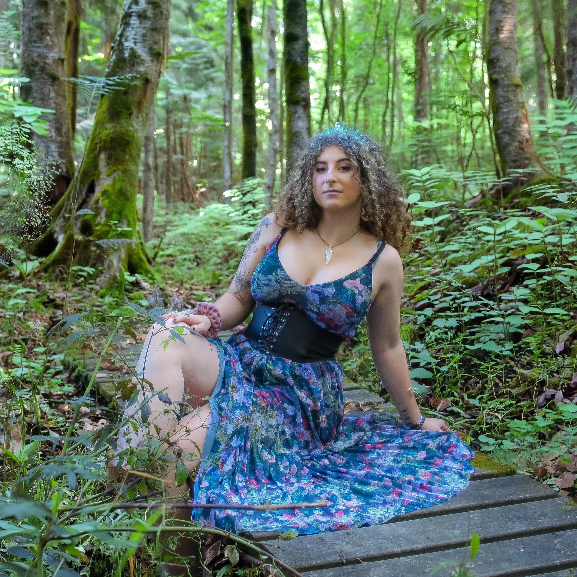 A young woman with curly hair and tattoos on her arm, wearing a floral dress, sitting on a wooden trail in a lush green forest with tall trees and dense foliage.