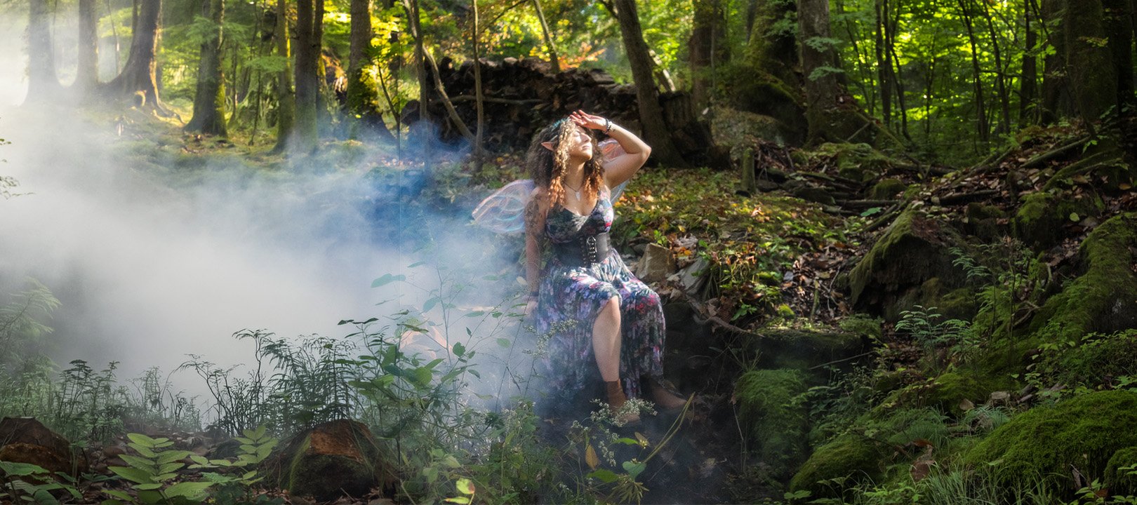 A woman with fairy wings in a forest, sitting on a log, shielding her eyes from sunlight, surrounded by lush greenery and moss-covered rocks, with mist or smoke nearby.