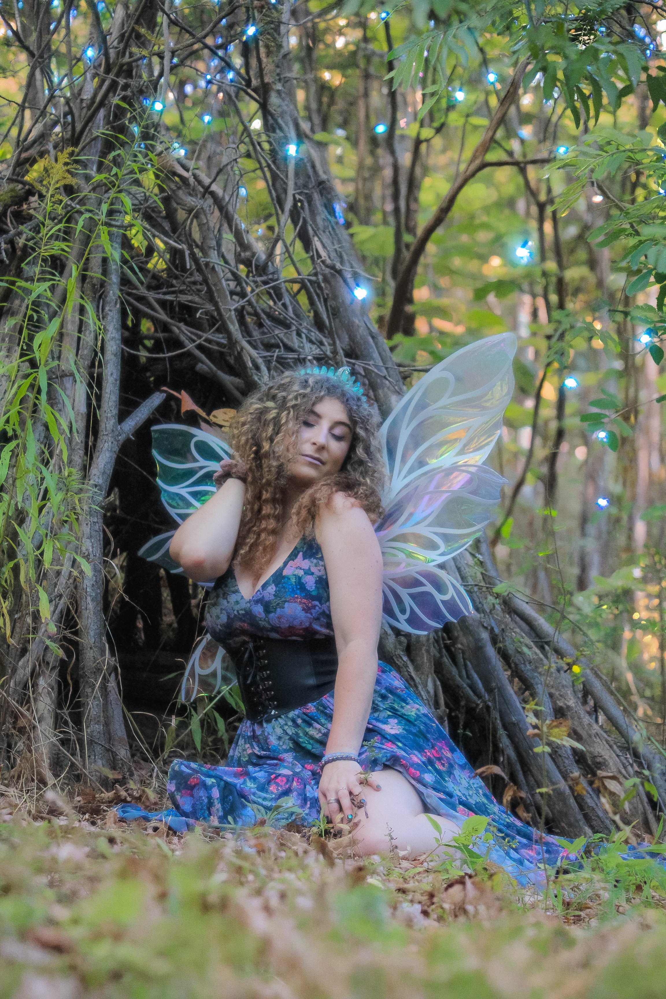 A woman dressed as a fairy with iridescent butterfly wings, a floral dress, and curly hair, sitting outdoors among trees with fairy lights and a natural setting in the background.