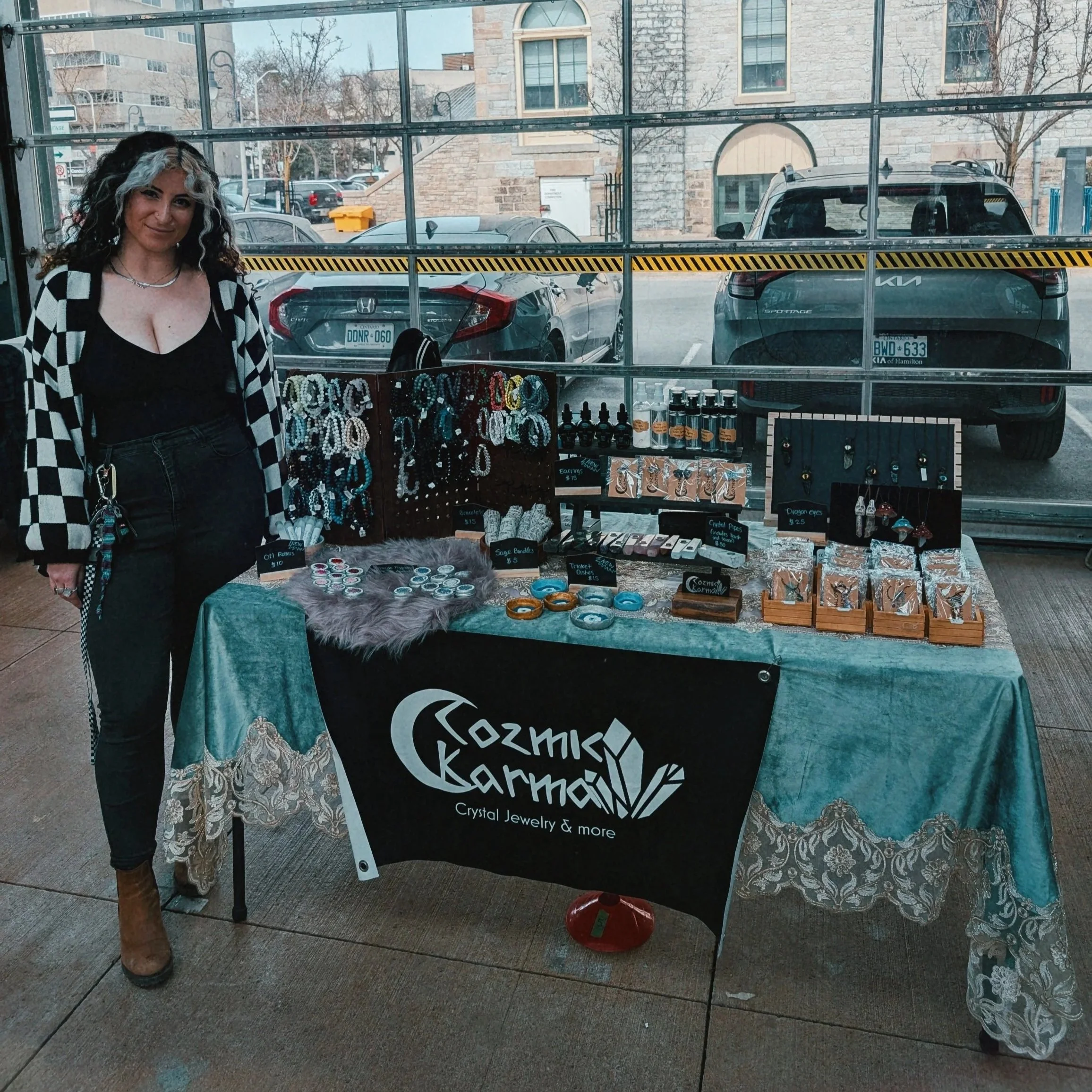 A woman with black and white curly hair stands in front of a jewelry booth at an outdoor market. The table is covered with a teal cloth and displays various jewelry items, including earrings, bracelets, with handwritten price tags. Behind her, a parking lot with cars and a stone building are visible through large windows.
