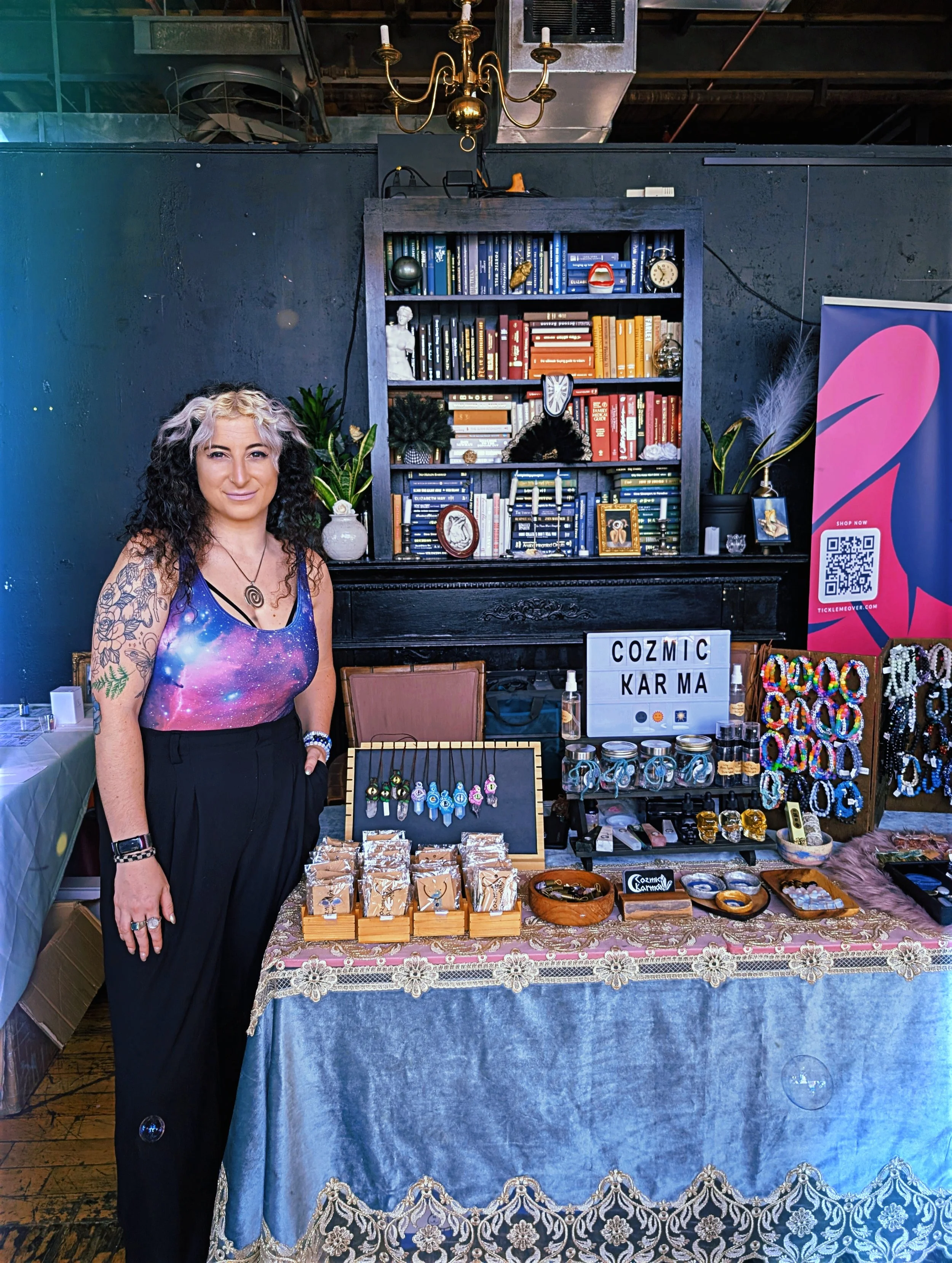 A woman with curly hair, wearing a galaxy-themed sleeveless top, stands next to a display table filled with jewelry and accessories at a booth. Behind her, a black bookshelf with colorful books and decorative items is against a dark wall. To the right, a pink banner with a QR code is visible. The tablecloth has floral lace trim, and various jewelry pieces are neatly arranged on the table.