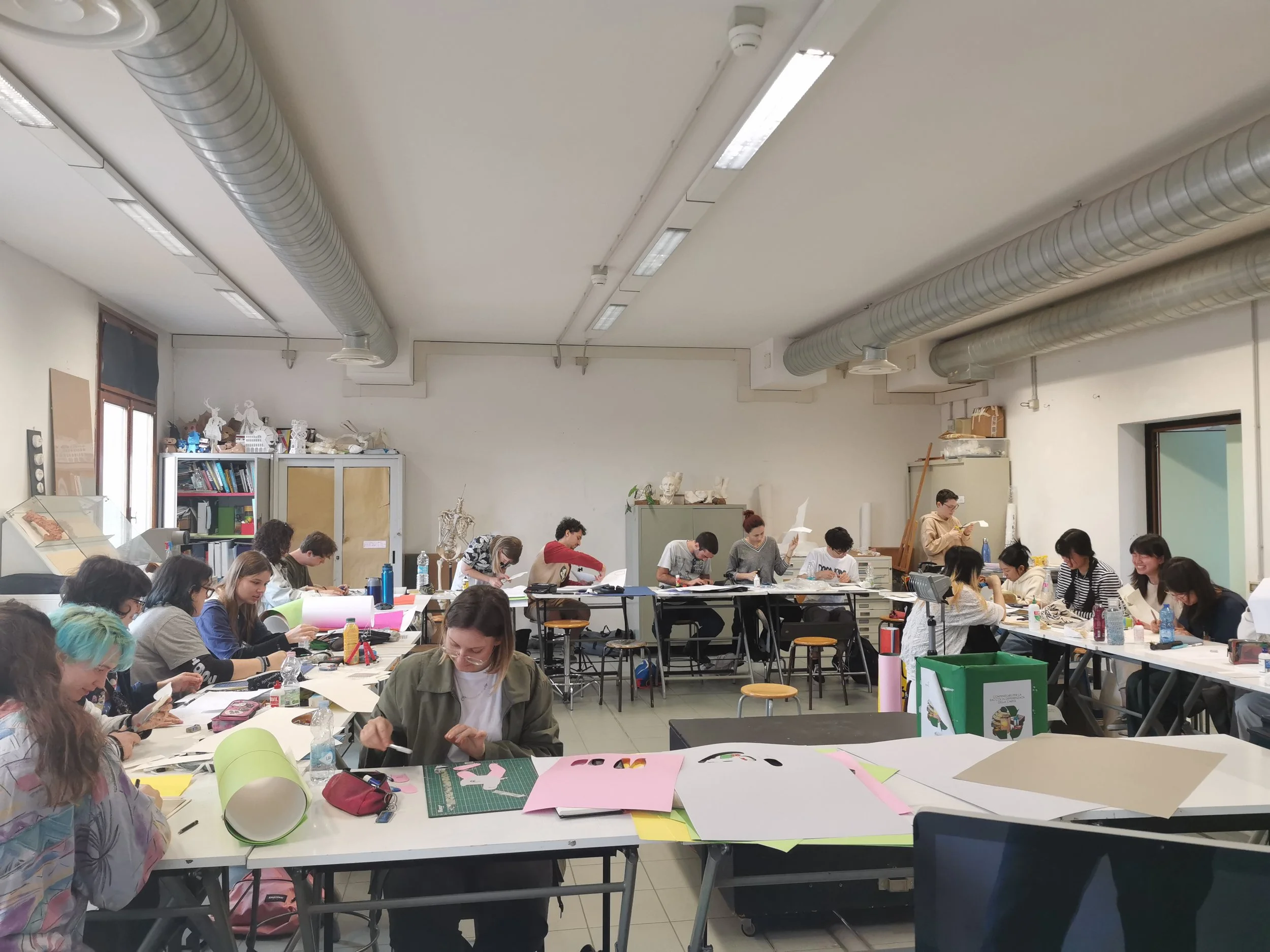 Un'aula con molte persone sedute a tavoli che lavorano su progetti artistici o artigianali, con materiali colorati e vari attrezzi. La stanza ha pareti bianche e soffitti con tubi di ventilazione visibili.