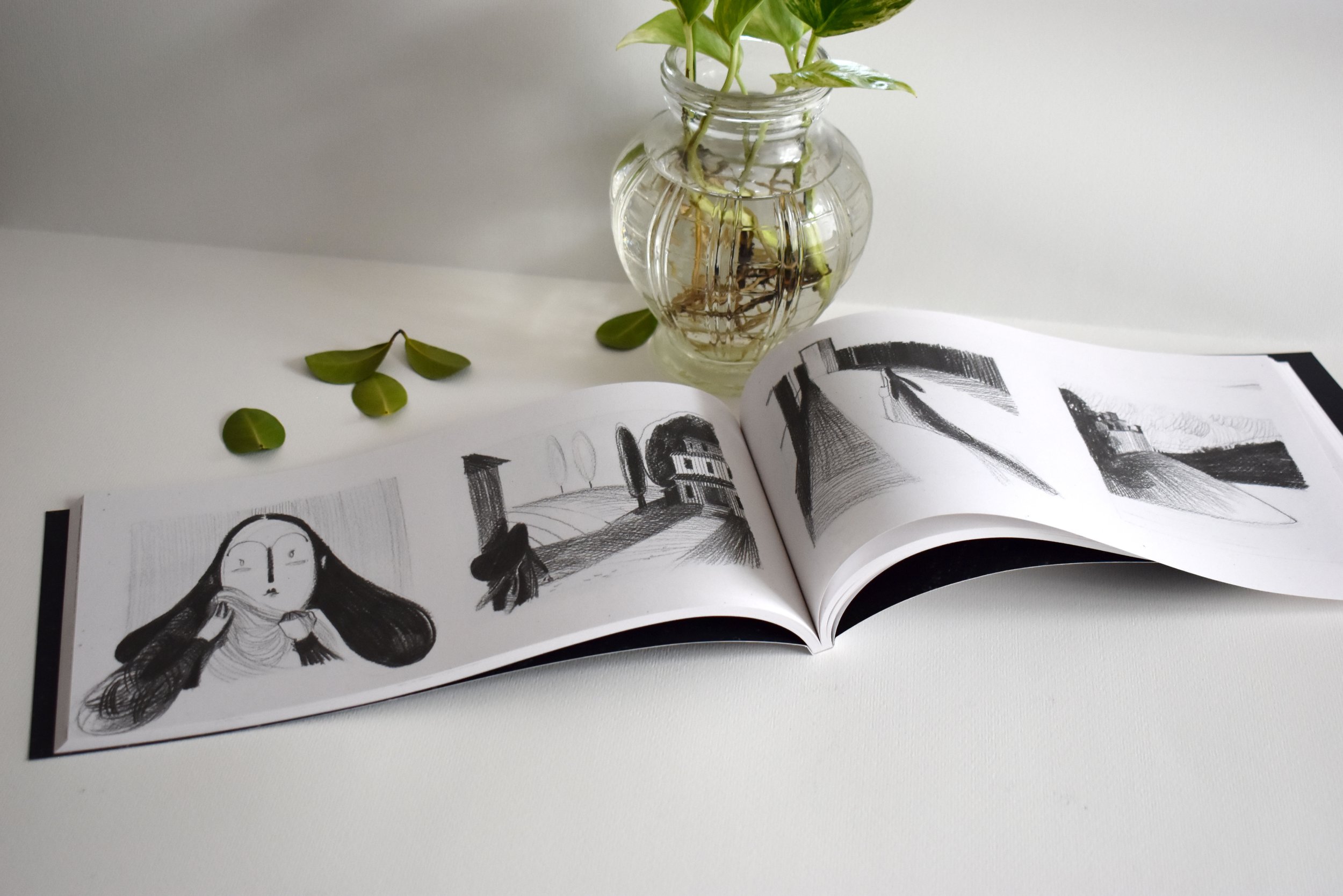 Un libro aperto con disegni in bianco e nero di un volto di donna, un paesaggio e una figura suoni, in posizione orizzontale, su una superficie bianca, con un barattolo di vetro con piante e radici e alcune foglie verdi sulla superficie.