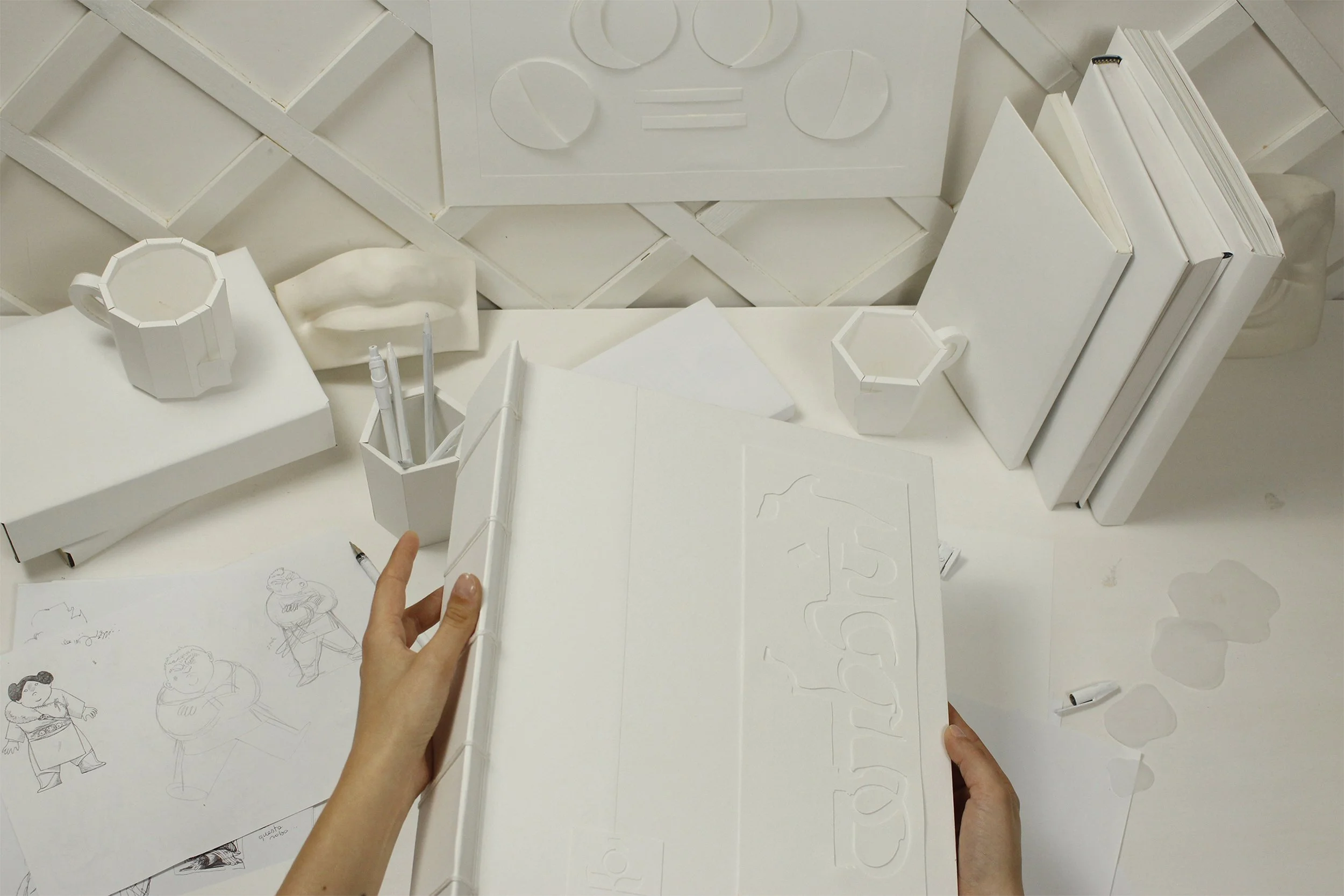 A person working on creating a 3D paper model in a workspace with white tools, books, sketches, and papers on a white table and background.