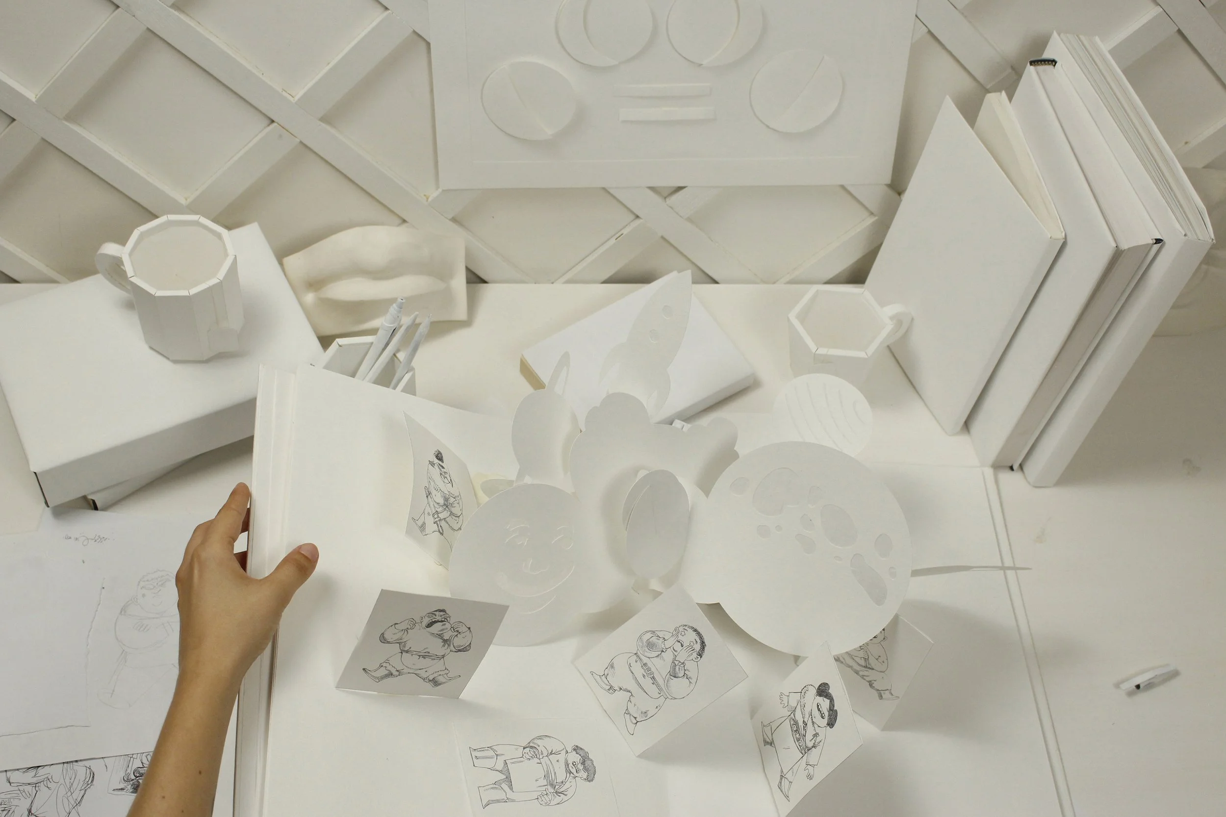 A person holding an open sketchbook with black-and-white character drawings. The table has paper cut-outs of faces, planets, and characters. There are white books, sketches, and paper objects on the table and some white objects on the wall behind. Th