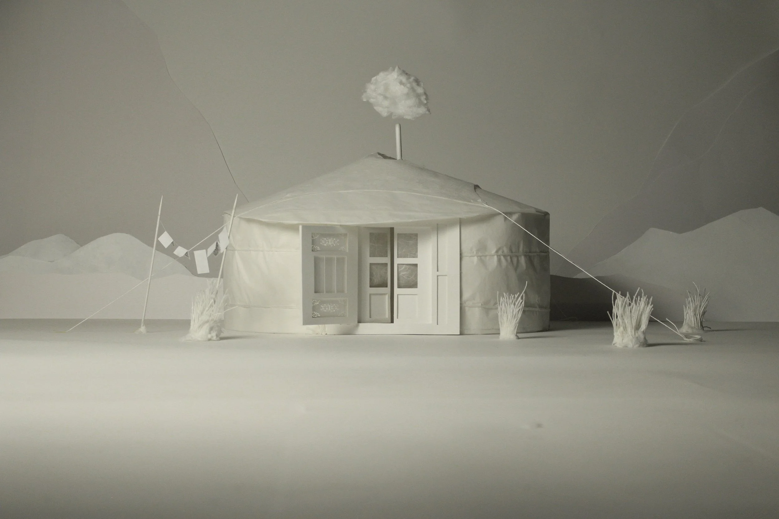 Modello di una casa di design in bianco, con una finestra e un'anta, su uno sfondo di montagne e un cielo grigio, con un albero di carta con un cerchio di nuvola sopra e alcuni cespugli di carta ai lati.